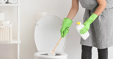 A person wearing a gray apron and light green gloves holds a bottle of cleaner and cleans a toilet.
