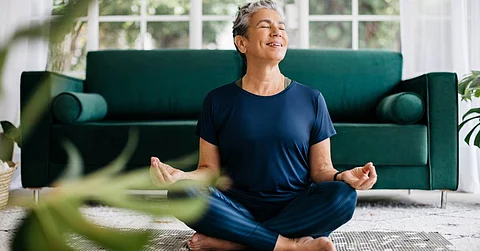 A woman in blue clothes closes her eyes and smiles as she sits on the floor with her legs crossed and meditates.