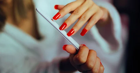 A close-up of a woman in a white bathrobe filing the ends of her bright-red acrylic nail manicure.