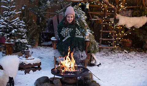 A woman in winter clothing sitting in a wooden arm chair as she smiles and looks down at the fire surrounded by snow.