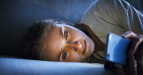 An upset woman lying on her couch, scrolling on her smartphone, which illuminates her face in a dark room.
