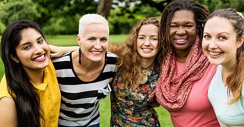 Five smiling women standing on a grassy field with their arms over each other's shoulders. One has a pink scarf.