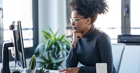 A woman wearing brown glasses and a gray turtleneck sits at a desk in an office, working on a computer.