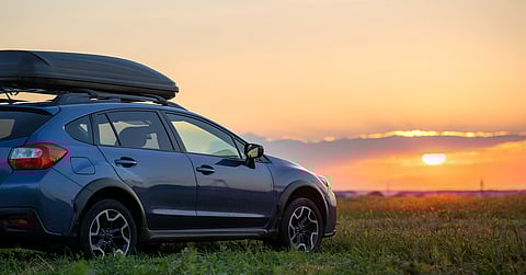An SUV with a cargo box on the roof of the vehicle. The car is parked on the grass in front of a sunset.