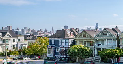 Multiple homes of different colors with small trees, with cars parked on the street and buildings in the background.