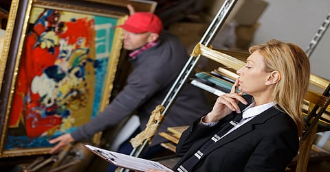 A woman in a black suit holds a clipboard and gazes at a painting held by a man in the background.