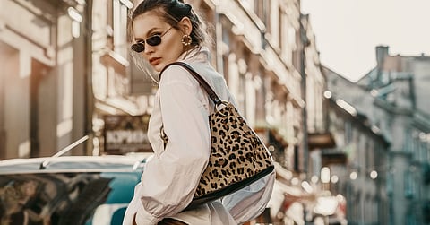 A stylish woman wearing sunglasses and a zebra print bag stand in the middle of the city street, next to cars.