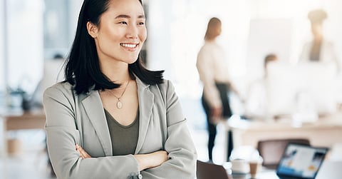 A woman wearing a light gray blazer smiles and stands with her arms folded. More workers are in the blurred background.
