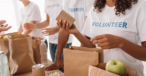 Volunteers in white shirts pack food donations into paper bags with canned goods and fresh produce on a table.