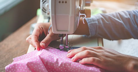 Hands guide pink polka-dot fabric under a sewing machine's presser foot. Soft light brightens one corner of the scene.