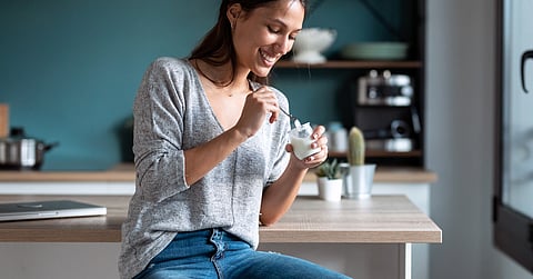 Woman sitting on kitchen counter smiles while eating yogurt from small glass jar with spoon in modern home.