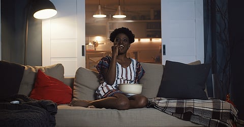 A smiling woman sits with her legs up on her couch and a bowl in her lap. She brings something up to her mouth.