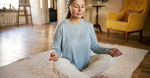 A woman is sitting cross-legged on a floor. She’s sitting on a carpet in the middle of a living room.