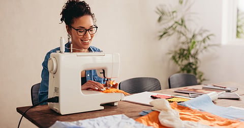 A smiling woman uses a sewing machine. Colorful fabric, sewing materials, and papers are spread across the workspace.