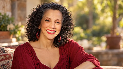 Dayna Farris Fisher smiling warmly in a red blouse, seated outdoors in soft golden light with a garden setting in the background.