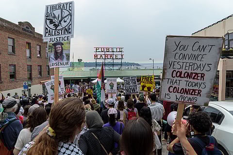 A large crowd of protesters marches near Pike Place Market by the waterfront, holding signs supporting Palestine. Prominent signs read "Liberate Palestine Justice," "Free Palestine," and "You Can't Convince Yesterday's Colonizer That Today's Colonizer Is Wrong." The iconic red "Public Market" sign is visible in the background, along with a view of the water and distant land.