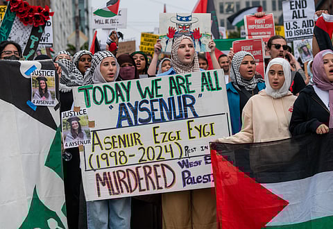 A group of protesters, mostly women wearing hijabs and keffiyehs, march while holding signs and flags. A central sign reads, "Today we are all Aysenur," and memorializes Aysenur Ezgi Eygi (1998-2024), stating she was murdered in the West Bank. Several signs call for justice, and one large Palestinian flag is visible.