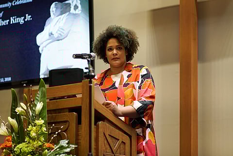 A woman with curly hair stands at a podium, holding papers and speaking into a microphone. She wears a vibrant, multicolored outfit. Behind her is a large screen displaying an image of Martin Luther King Jr. and text referencing a community celebration in his honor.