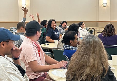 A group of people sits around tables during a watch party. Several individuals are smiling and laughing, while others raise their hands in participation. One person types on a laptop, and cups and papers are scattered across the tables.