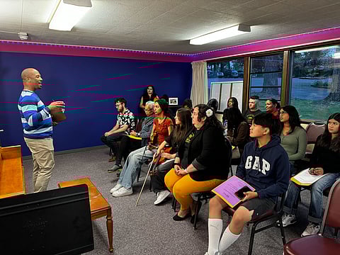 A man in a striped sweater stands at the front of a room, addressing a seated group of diverse individuals. Some hold musical instruments, like a violin and guitar, while others have papers or notebooks.