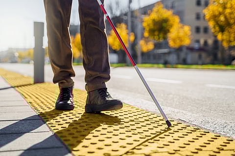 A person using a white cane walks along a tactile yellow surface with raised bumps at a sidewalk's edge. The cane is tapping the ground for guidance. The scene takes place outdoors on a sunny day, with trees and buildings blurred in the background.