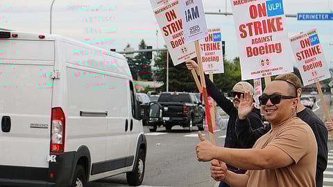 A group of smiling workers holds signs that read "On Strike Against Boeing" while walking on the side of a street. One man in the foreground wearing sunglasses gives a thumbs-up. Vehicles, including a white van, pass by on the road. The strike is organized by the Machinists Union (IAM District Lodge 751).