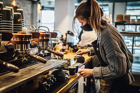 A barista wearing a mask and apron pours steamed milk into a cup of coffee at an espresso machine in a cozy café. Stacks of branded coffee cups are visible on the counter, and the warm, inviting atmosphere of the coffee shop is evident, with soft lighting and equipment in the background.
