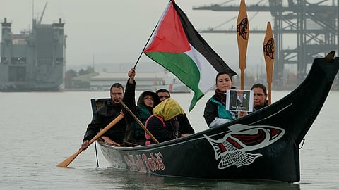 A group of people paddle a traditional canoe on a body of water, holding a Palestinian flag and a "Free Palestine" sign. The canoe features indigenous artwork, including a fish design on the side. The scene takes place near industrial structures and cranes, suggesting a protest or demonstration advocating for Palestinian solidarity.