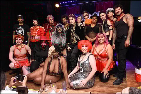 A diverse group of performers pose together on stage, wearing a variety of bold and creative costumes. Outfits include military-inspired attire, glittery dresses, leather harnesses, and vibrant wigs. Some individuals sport face paint or crowns.