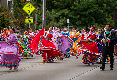 A group of women in colorful folkloric dresses dance in unison during a parade, twirling their vibrant skirts. The dresses are red, purple, green, and yellow with colorful ribbons, creating a lively display. A man in a black charro suit walks beside them, smiling.