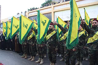 A group of uniformed Hezbollah militants stands in formation, holding yellow flags with green insignias. They wear camouflage uniforms with matching berets and sashes, and some extend their right arms in a salute.