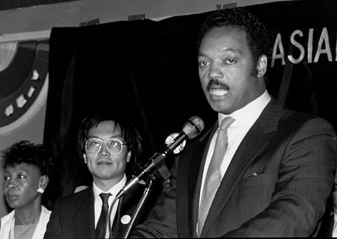 A black-and-white photo of a Rev. Jesse Jackson speaking at a microphone, wearing a suit and tie. Two individuals, an Asian man in glasses and a Black woman, stand behind him, listening attentively. The backdrop partially shows the word "ASIAN,"