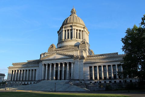 The Washington State Capitol building in Olympia, a grand structure with a large dome and neoclassical design. The facade features tall columns, wide stone steps, and intricate architectural details. The building is lit by sunlight under a clear blue sky, with trees partially visible to the right.