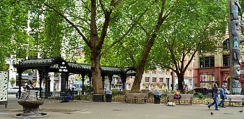 A peaceful urban park scene featuring Pioneer Square in Seattle. Large trees with lush green leaves provide shade, and a historic pergola stands in the center. A totem pole is visible on the right, and a few people are sitting or walking nearby.