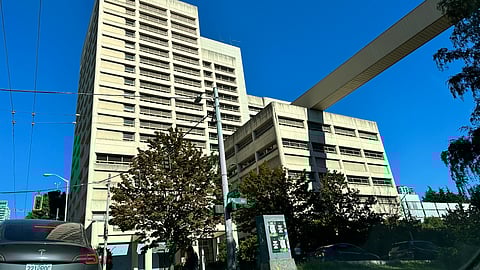 A large, beige, concrete building stands tall against a clear blue sky. The structure features many narrow windows and a pedestrian skybridge connecting it to another part of the complex. Below, trees partially obscure the lower floors.