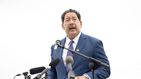 A man in a blue suit with a dark tie and lapel pin speaks at an outdoor event, standing behind a podium with multiple microphones. He looks to the side while addressing the audience.