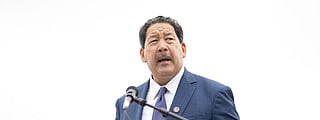 A man in a blue suit with a dark tie and lapel pin speaks at an outdoor event, standing behind a podium with multiple microphones. He looks to the side while addressing the audience.