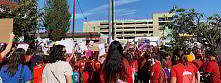 A large crowd gathered outdoors around a flagpole with the American flag, many wearing red shirts and holding signs. The event appears to be a demonstration or rally. Buildings, trees, and power lines are visible in the background.