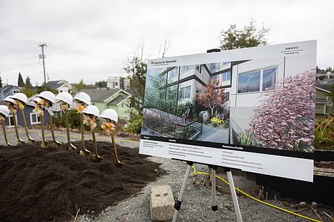 A row of golden shovels with white hard hats, each adorned with orange ribbons, is stuck into a mound of dirt at a groundbreaking ceremony. Next to them, a large display board on a tripod shows a rendering of a future housing project by Habitat for Humanity, featuring details about unit sizes and residence profiles.
