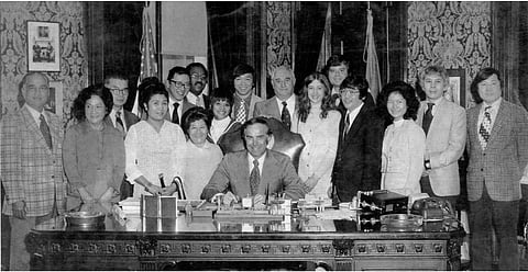 The late Gov. Dan Evans with the first Washington State Commission on Asian Pacific American Affairs (CAPAA) in 1972. He sits at a desk in the center of the commission members, who are standing around him and the desk and smiling at the camera.