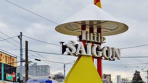 A close-up of a sign for "Little Saigon," featuring bold white letters mounted on a yellow, cone-shaped structure with a conical white roof resembling a traditional Vietnamese hat. Power lines crisscross the sky in the background, and nearby buildings are partially visible under a cloudy sky.