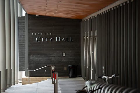 A view of the entrance to Seattle City Hall, featuring dark tiled walls with "Seattle City Hall" prominently displayed in silver letters. The area is partially enclosed, with a modern design that includes metal columns, glass doors, and a wooden ceiling. A bicycle rack is visible in the foreground.
