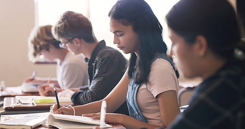 A group of students sits in a classroom, focused on their work. The student in the foreground, a young woman in a pink shirt and overalls, writes in a notebook. Others around her, slightly blurred, are also engaged in writing or studying.