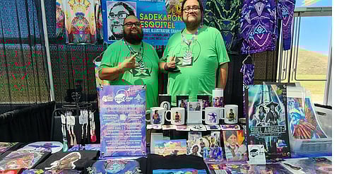 Two men stand behind a booth at an event, both wearing matching green t-shirts. Their booth displays a variety of artwork, including posters, mugs, and t-shirts featuring colorful designs. A banner behind them promotes "Sadekaronhes Esquivel" as an artist, illustrator, and graphic designer, with details about their services.