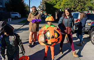 A group of people trick-or-treating on a sunny day. A child in the center is dressed in an inflatable pumpkin costume, smiling and holding a paper bag. A man behind carries candy, while a woman walks beside them, also smiling.