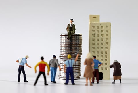 Numerous miniature people gathered in front of miniature buildings and piles of coins.