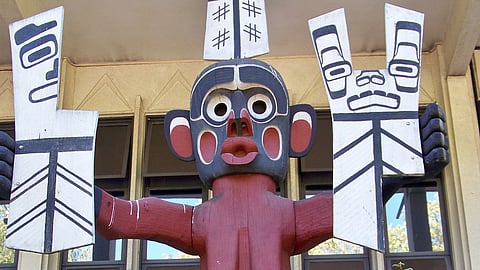 A wooden Indigenous totem pole with a stylized figure featuring bold black, white, and red paint. The figure has a rounded face, wide eyes, and open mouth, holding two large rectangular objects with black geometric patterns. The totem is displayed in front of a building with beige walls and large windows.