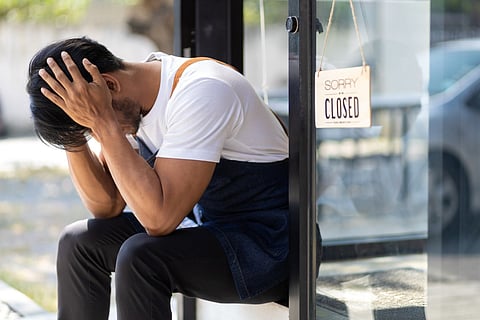 A man wearing an apron sits outside a storefront with his head in his hands, appearing stressed. A "Sorry, we are CLOSED" sign hangs on the door next to him. The setting suggests a business closure, and the man's posture reflects frustration or exhaustion.