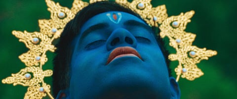 A close-up of a person with blue-painted skin and closed eyes, wearing an ornate golden headdress. The person has a red and white marking on their forehead and appears to be in a calm or meditative state.