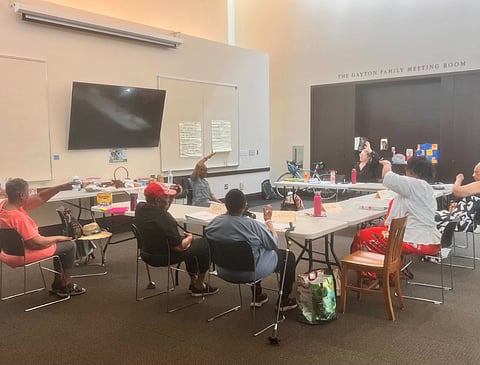 A Matter of Balance workshop held at the Douglass-Truth Branch in partnership with The Seattle Public Library and funded by Pacific Hospital PDA. People sit around tables arranged in a U-shape, with a presenter at the center. They are all participating in an activity, each one raising an arm above their head.